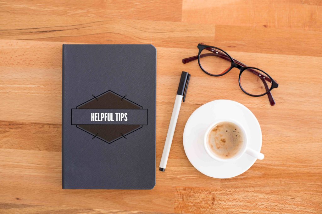 Black journal that says helpful tips on it next to a pen, cup of coffee, and glasses atop a wooded table